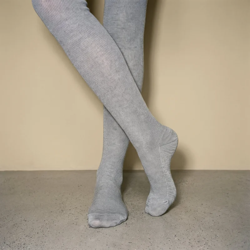 Both feet of a female model wearing grey ribbed tights are shown from an angled front view with the ankles crossed. A sand dune studio-wall backdrop frames the composition.