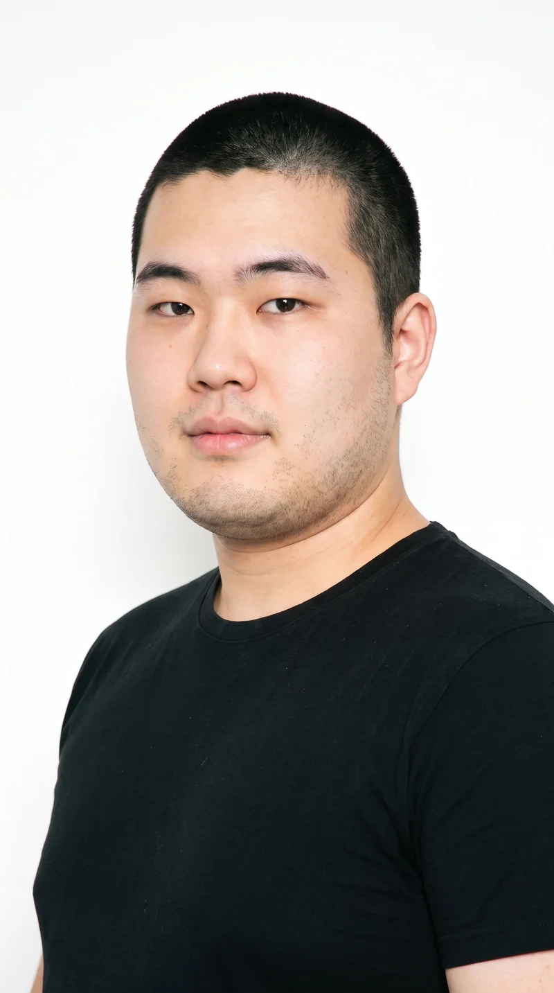 A 22-year-old Japanese man with East Asian facial features, light porcelain skin with a warm yellow undertone, and a round face defined by soft, full contours and few angles. He has thick eyebrows, very dark near-black eyes, and medium lips. His black hair is clipped very short and evenly in a buzz cut, and very short facial-hair growth creates a faint, even shadow across the beard area.