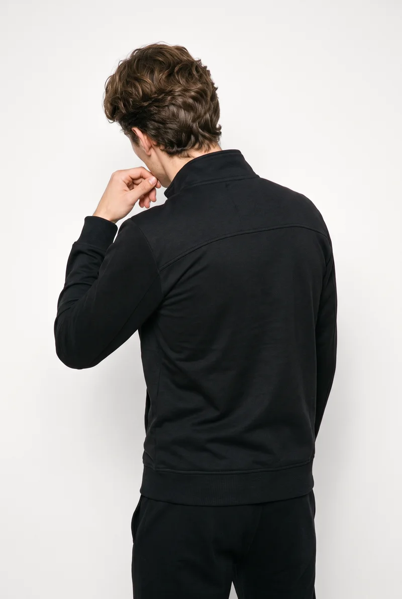 A 21-year-old Caucasian man wears a black tracksuit in an upper-body back view, with one hand at his chin. A Chantilly Lace studio-wall backdrop frames the composition.