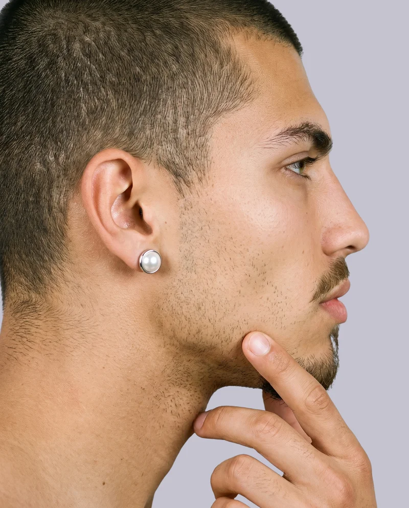 A side view of an ear wearing a men's pearl stud earring, with a finger positioned at the jaw. The subject is framed closely around the ear against a flat lavender-grey backdrop.