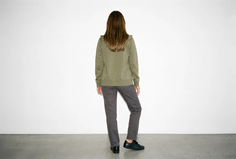 A 25-year-old Hispanic woman stands in a relaxed pose, shown from behind wearing a women's olive zip-up hoodie. The garment is framed in an environmental view against a pure white studio-wall backdrop.