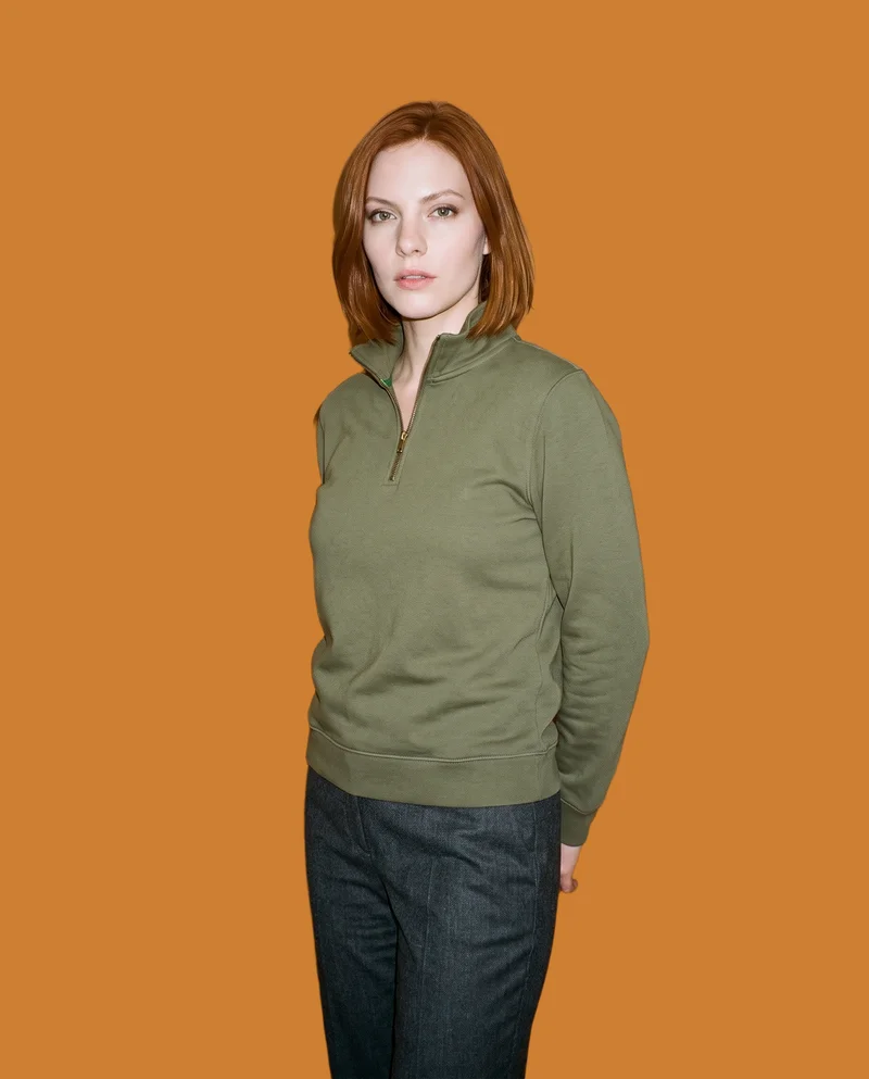 A 26-year-old Caucasian woman wears an olive quarter-zip top in a three-quarter-length view. She stands at a front three-quarter angle with her hands behind her back and a serious expression. A flat bronze backdrop frames the studio-cutout composition.