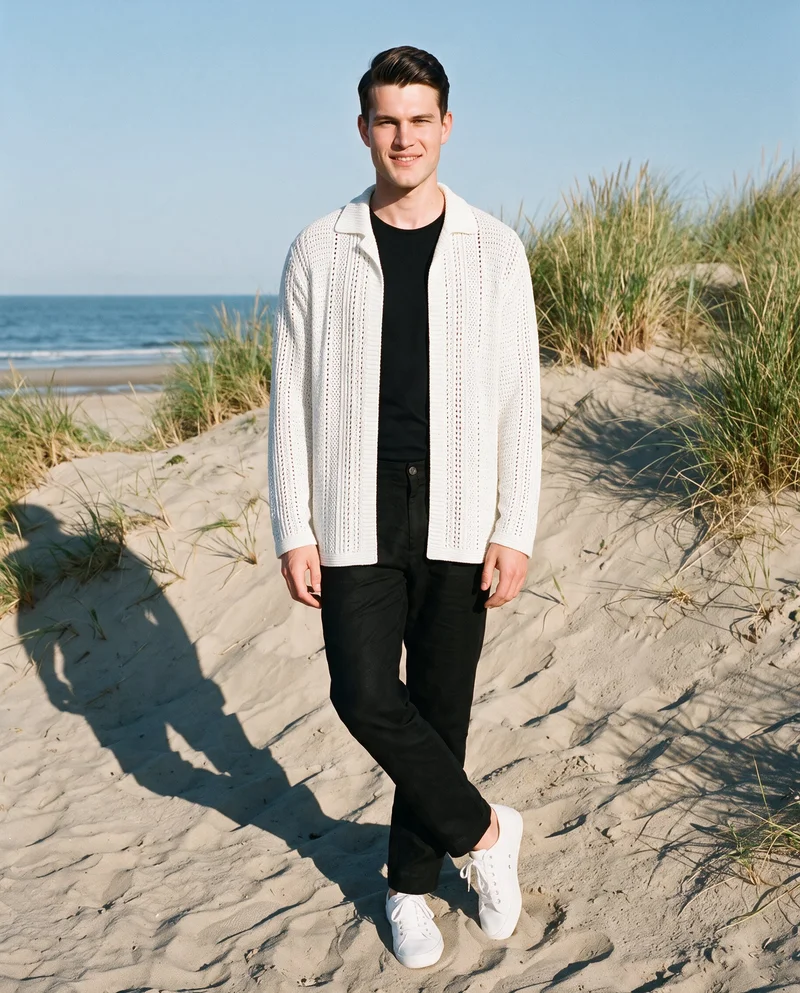 A 19-year-old Caucasian man wears a white crochet cover-up in a full-body front view, standing with his ankles crossed and smiling. A Martha's Vineyard beach backdrop is visible behind him.