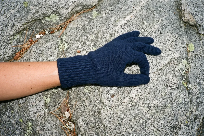 A close-up top view shows a hand wearing a navy ribbed glove, with the thumb positioned toward the fingers. The hand and wrist are framed against an Acadia coastal rocks backdrop.