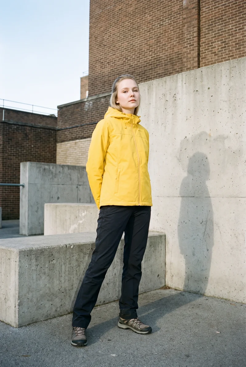 A 20-year-old Slavic woman wears a yellow waterproof jacket, shown full body from a front three-quarter angle with her hands behind her back and a confident expression. Parkour-style urban obstacles form the backdrop.