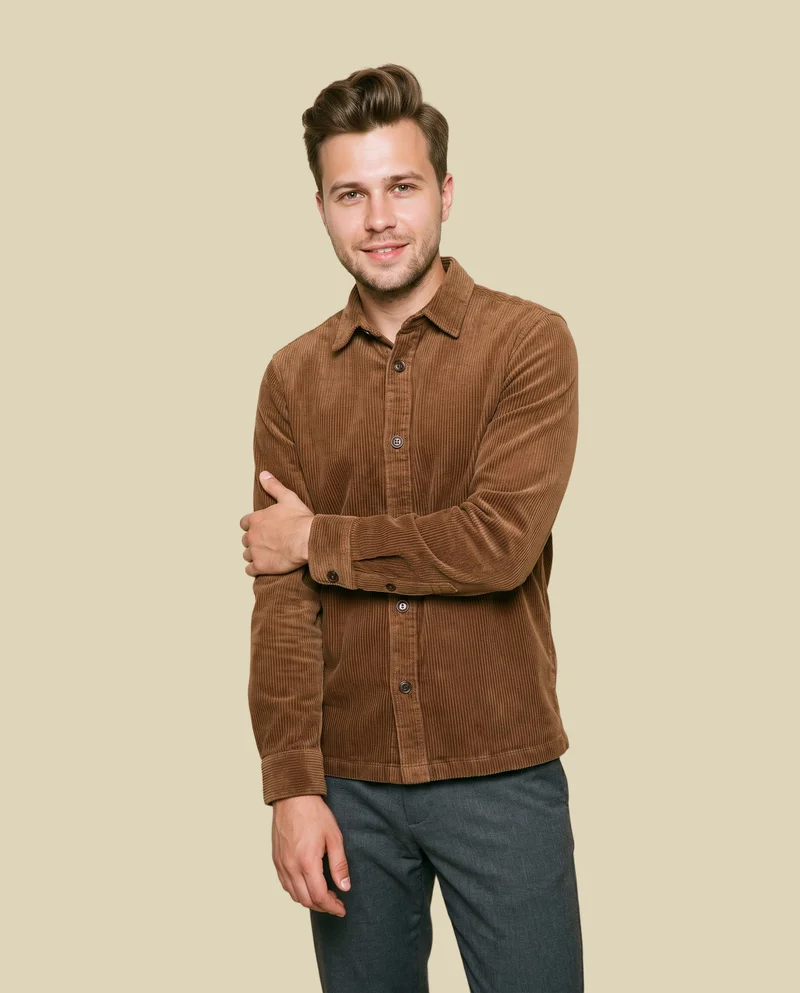 A 27-year-old Slavic man wears a brown corduroy overshirt in a three-quarter-length view from a front three-quarter angle. He is smiling and holding one arm, with a wheat-field flat-color backdrop.