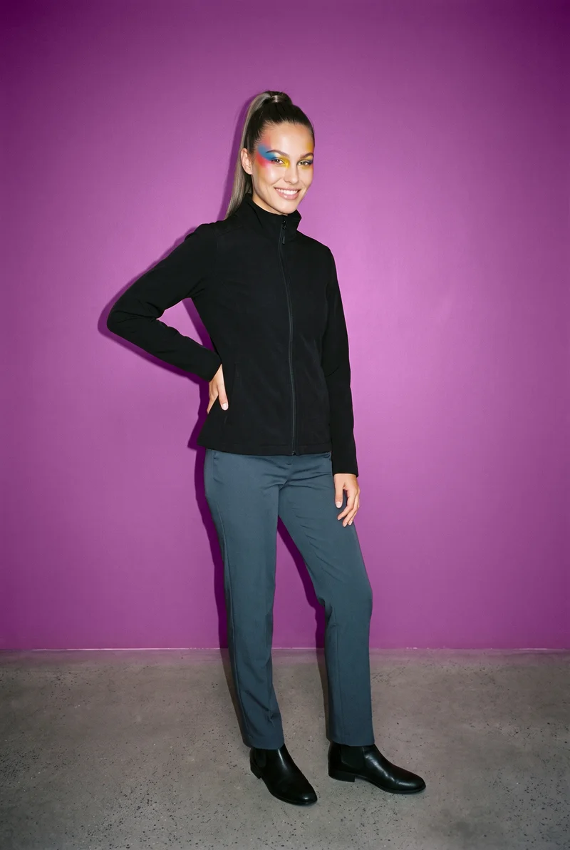A 23-year-old Caucasian woman models a women's black softshell jacket in an environmental front three-quarter view. She is smiling with one hand on her lower back, against a plum studio-wall backdrop.
