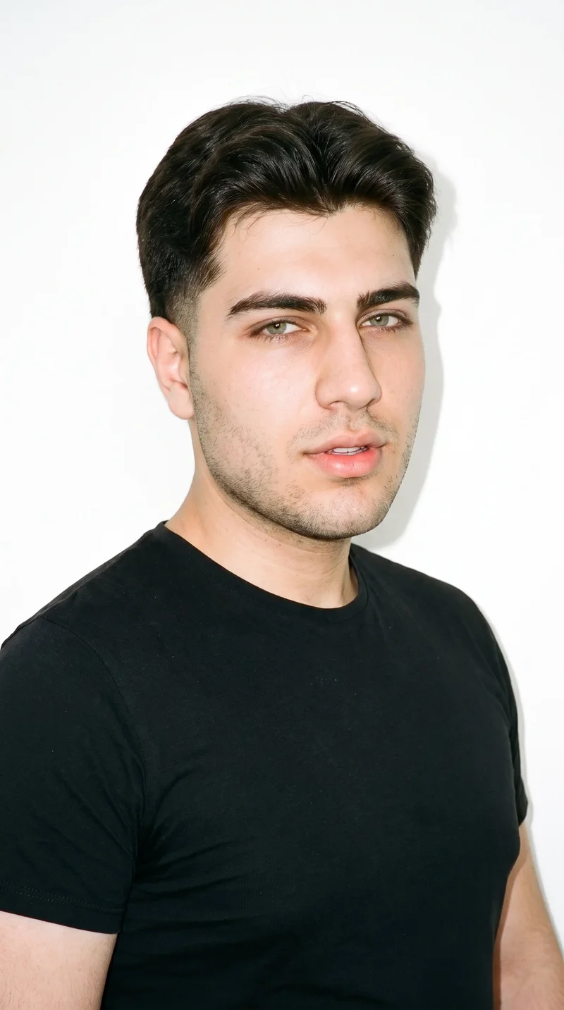 A 19-year-old Turkish man with lightly tanned skin and an olive-green undertone. His square face has a broad forehead and a strong, defined jaw, with thick eyebrows, green eyes, and medium-sized lips. His dark-brown hair is styled in a two-block cut, with short sides and back contrasted by a longer, fuller top. Fine, closely trimmed stubble lightly covers the beard area.