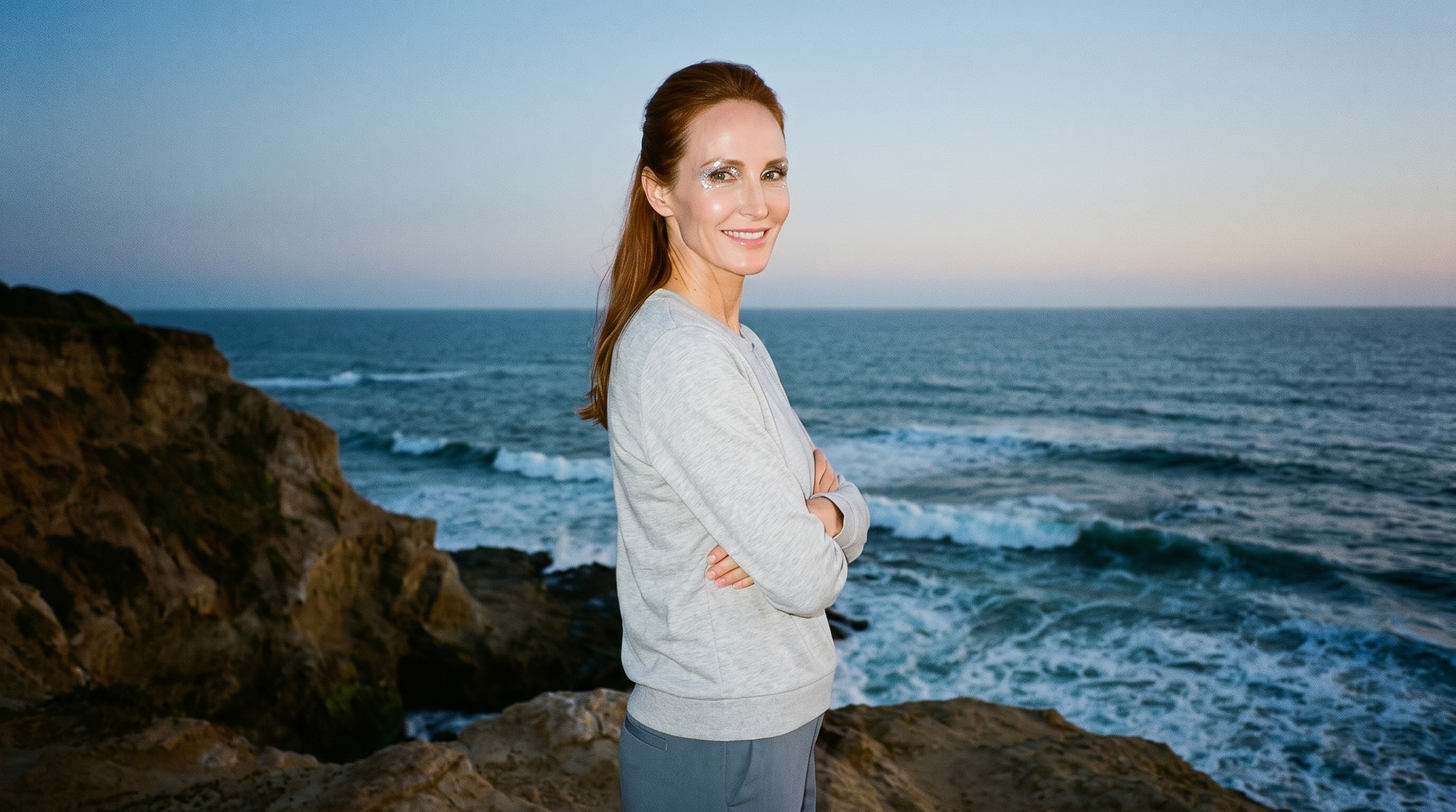 A 54-year-old Caucasian woman wears a women's grey tracksuit top in a three-quarter-length side view. She is smiling and holding herself in a self-hug pose, with a Coastal Cliff Pacific Waves backdrop.