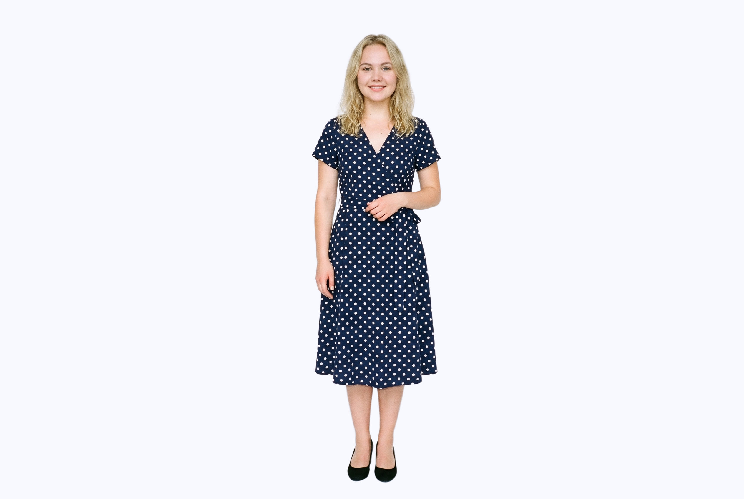 A 22-year-old Slavic woman wears a polka-dot wrap dress in a full-body front view. She is smiling with one foot positioned forward against a flat porcelain-colored backdrop.
