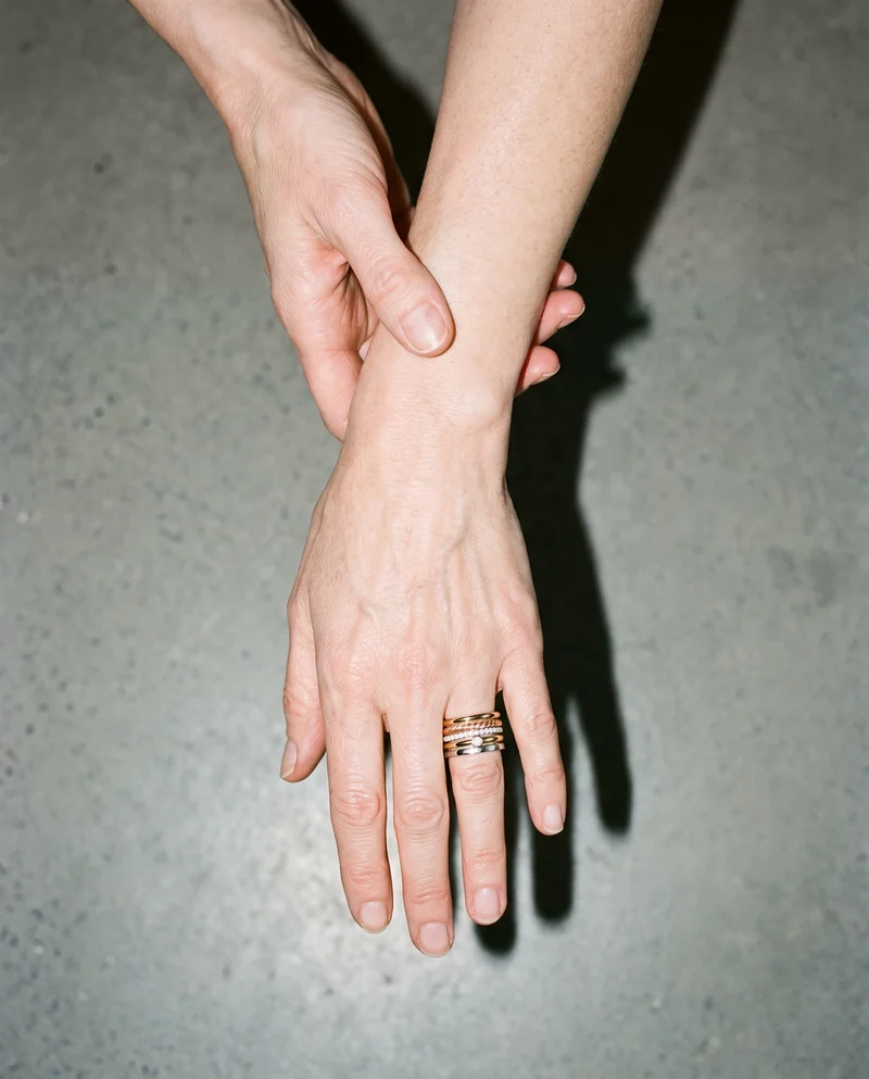 A hand and wrist are shown from above, with a women's mixed-metal stacking ring set visible on the fingers as the hand touches the wrist. The alabaster studio-wall backdrop fills the background.