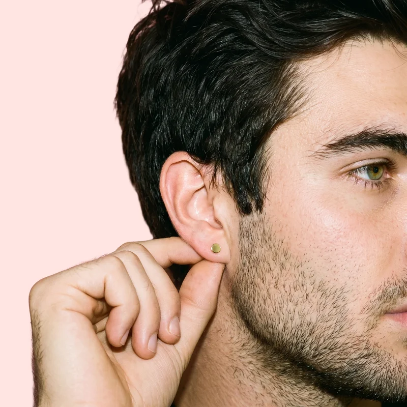 A man's ear is shown in side view with a finger touching the earlobe and a gold stud earring visible. The image uses a flat Misty Rose backdrop.