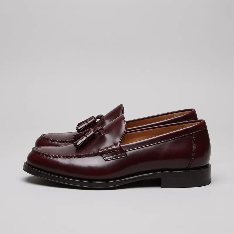 A pair of burgundy low-profile slip-on tassel loafers is shown in a side-facing view. The shoes have rounded moc toes, raised apron stitching, saddle straps across the vamps, tassel ornaments with knotted side ties, stitched side panels, low stacked block heels, visible sole edges, and vertical heel counter seams.
