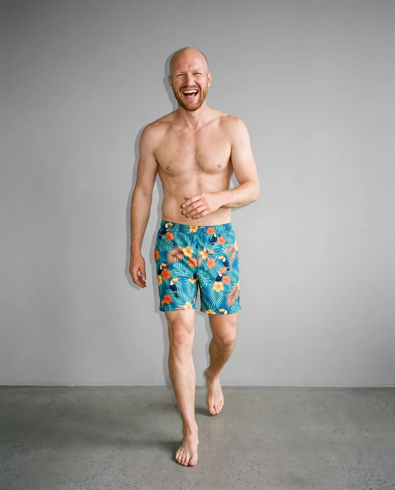 A 35-year-old Caucasian man is shown full body from the front while walking and laughing, wearing tropical print board shorts. A Pebble Grey studio-wall backdrop fills the background.