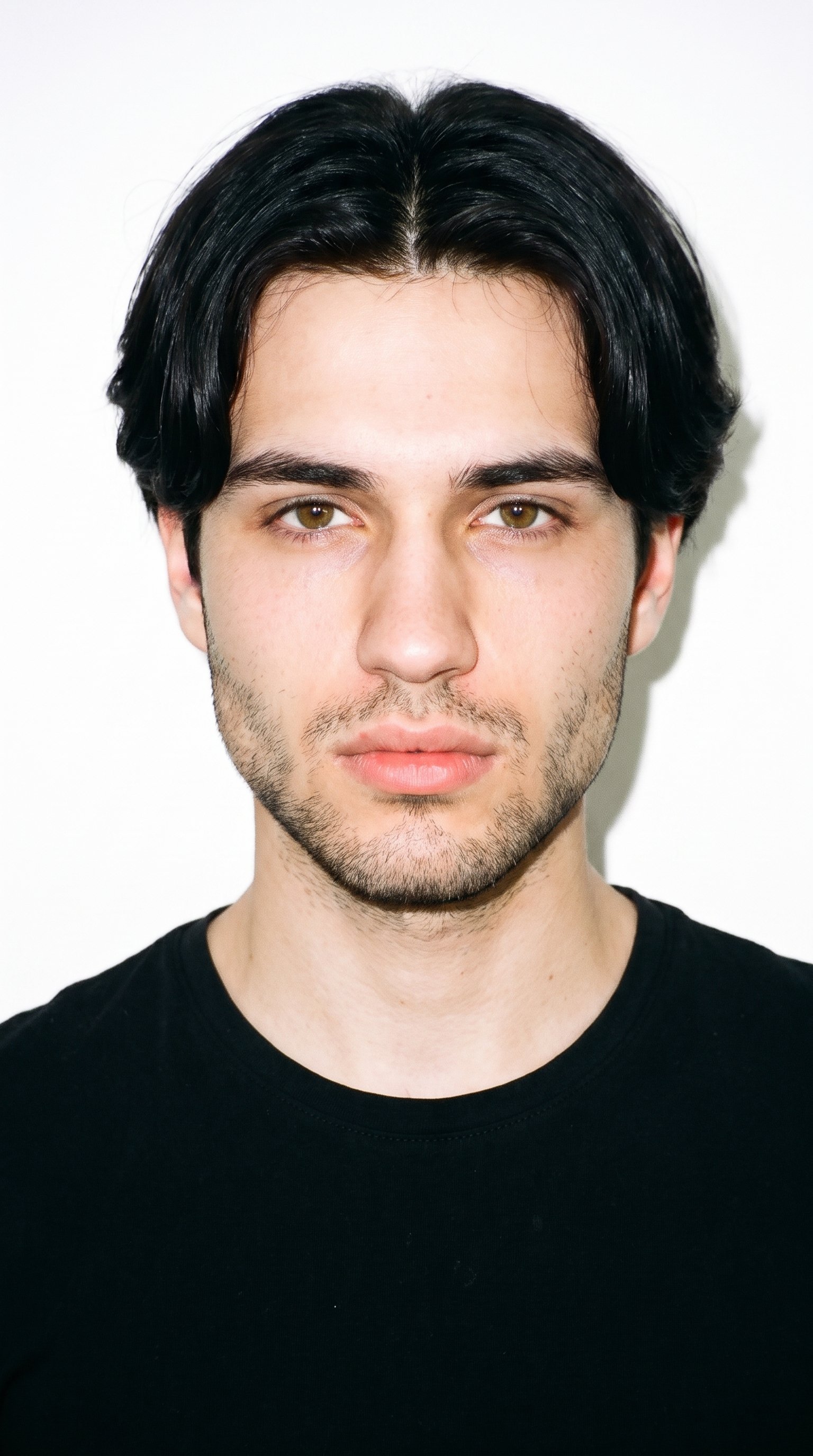 An 18-year-old Slavic male with fair skin and a neutral undertone has a triangle-shaped face, with a wide jaw narrowing toward the forehead and emphasis on the lower face. His eyebrows are thin, and his hazel eyes contain green-gold flecks. He has full lips and black hair divided at the centre, falling evenly to both sides. Dense heavy stubble forms a dark, defined layer across the beard area.