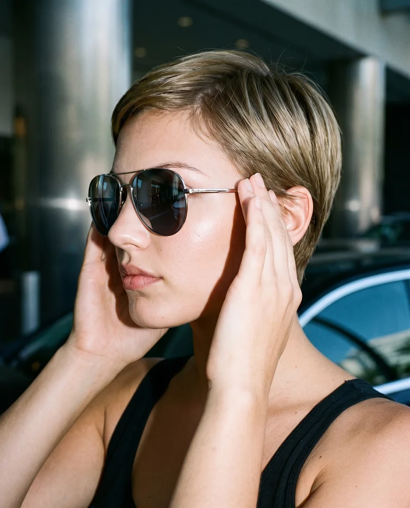 A 24-year-old Caucasian woman is shown in a head-framed side view wearing women's mirrored sunglasses. Her hands are at her head, and she has a serious facial expression. A valet stand serves as the backdrop.