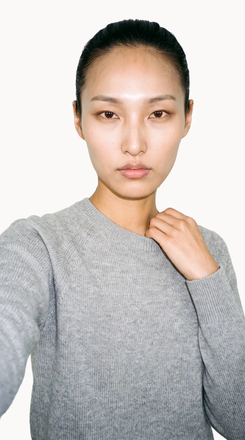 A 27-year-old Korean woman wears a grey cashmere knit in a front-facing selfie-style view, with one hand at her collarbone and a natural expression. A Chantilly Lace flat-color backdrop fills the background.