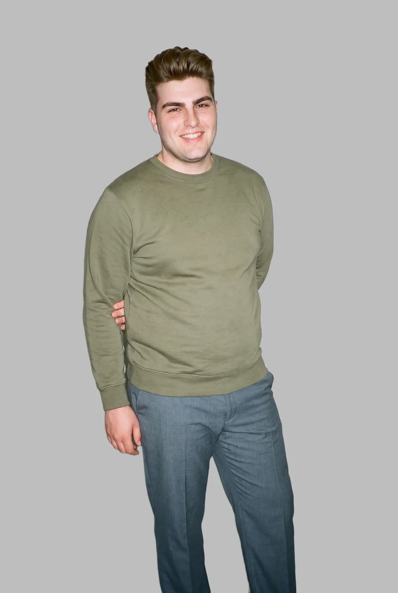 An 18-year-old Caucasian male model wears a men's olive crewneck sweatshirt in a three-quarter-length front view. He is smiling with one elbow positioned behind his back against a dove grey flat-color backdrop.