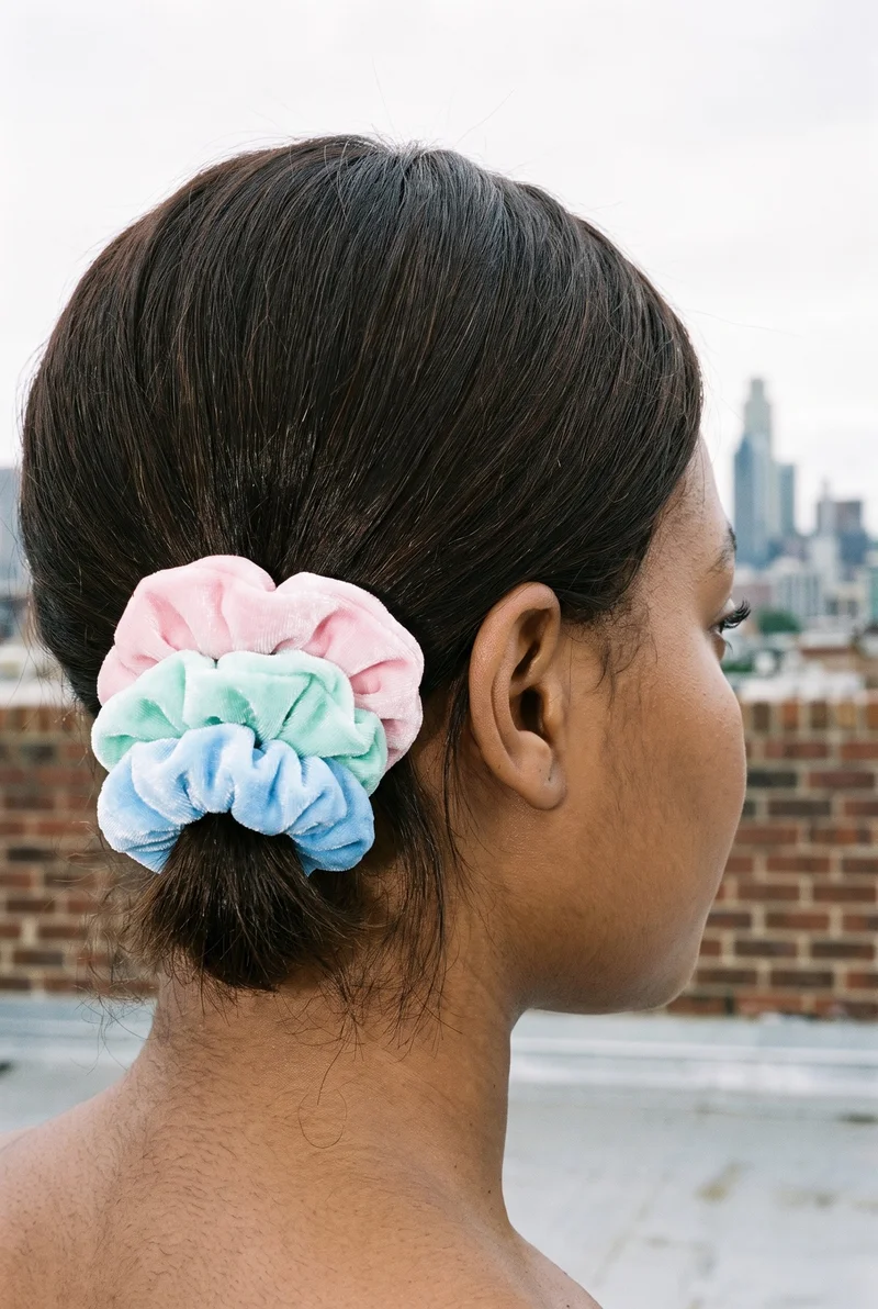 A 28-year-old African woman is shown from an angled rear view with her head level, wearing the Pastel Scrunchie Set. The head is framed against a warehouse rooftop skyline backdrop.