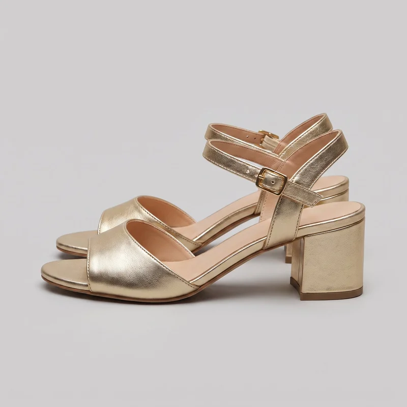 A pair of gold open-toe heeled sandals is shown in a side three-quarter view. Each sandal has a wide forefoot band, an exposed footbed, an open waist and back, a vertical heel strap, and an adjustable ankle strap with a rectangular buckle. The sandals have mid-height block heels, narrow edge piping, stitched strap borders, a thin outsole edge, and layered heel bases.