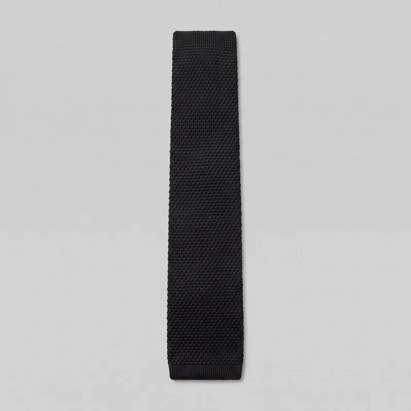 A complete flat view of a black knitted necktie with a long, narrow rectangular shape, straight parallel sides, and squared top and bottom ends. The surface has a uniform dense textured stitch pattern, with slightly denser ribbing at the end bands and softly rounded edges from the knit construction.