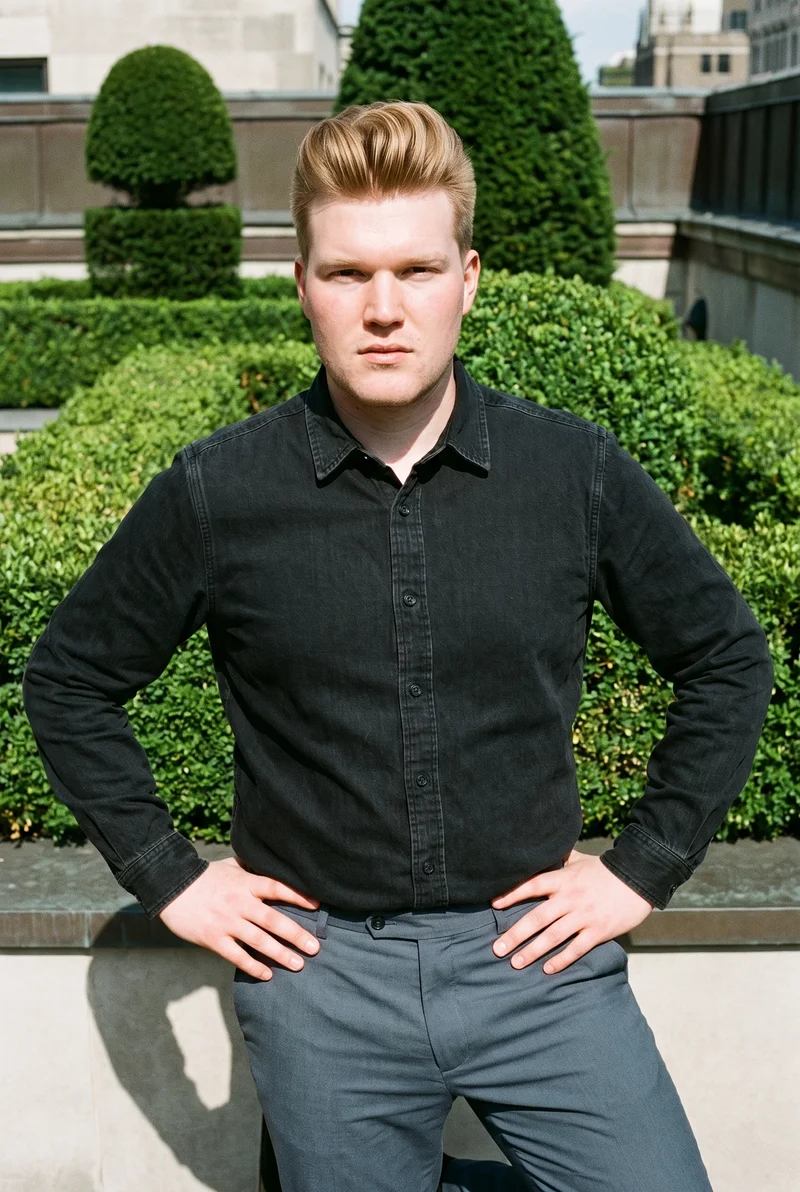 A 23-year-old Caucasian man wears a black denim shirt in a three-quarter-length front view, standing with his hands on his waist and a confident expression. A formal rooftop garden serves as the backdrop.