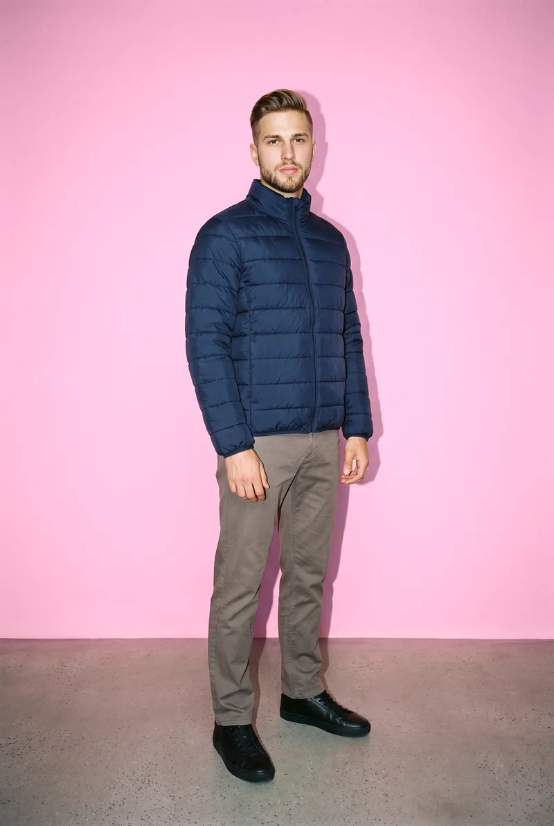 A 22-year-old Caucasian male model wears a men's navy padded jacket in a full-body view from the front three-quarter angle. He stands with his hands in front and a confident expression against a Cotton Candy studio-wall backdrop.