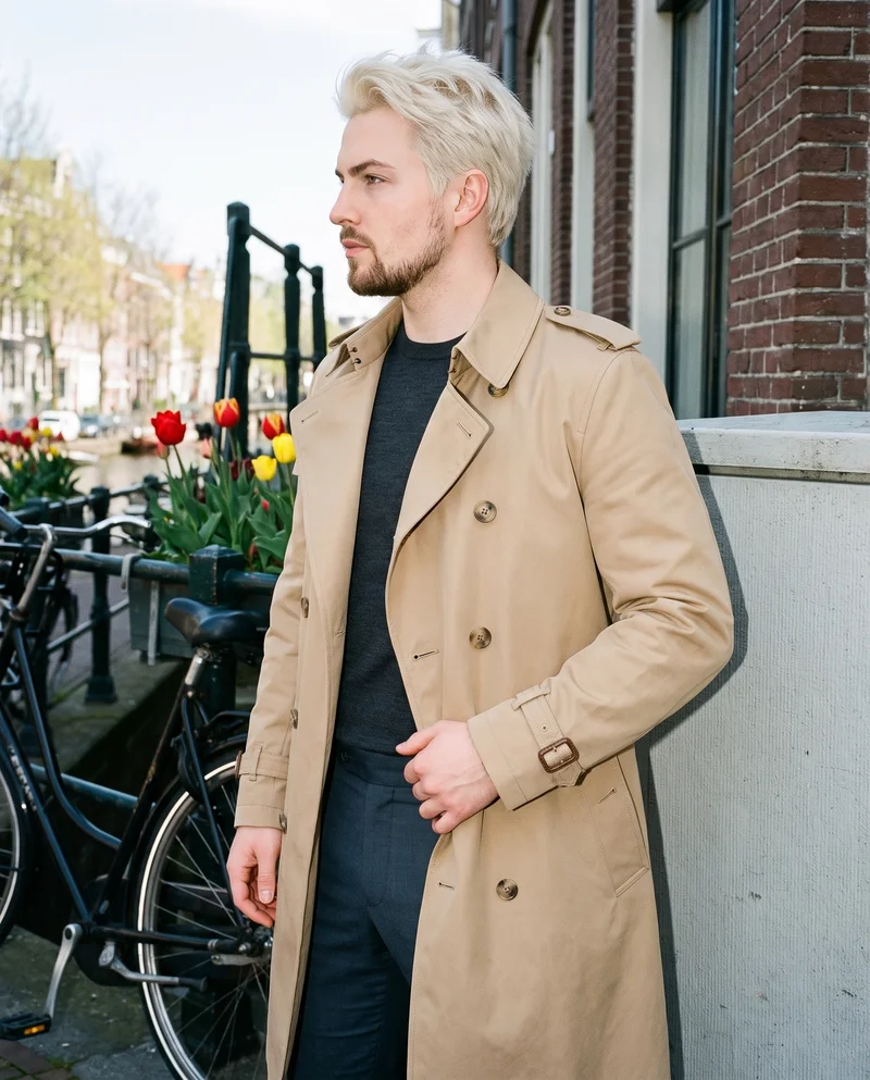 A 23-year-old Caucasian man wears a men's khaki belted trench coat in a three-quarter-length side view, standing with his weight shifted back and a neutral expression. An Amsterdam canal house forms the backdrop.