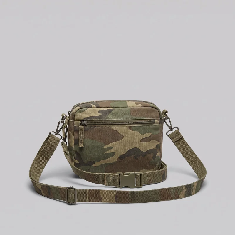 A rectangular camouflage crossbody bag is shown alone in a front view, with its strap spread around the bag. The structured boxy body has rounded upper corners, piped edges, a top zipper, and a horizontal front zipper pocket. Snap hooks, D-ring hardware, a strap slider adjuster, and a side-release buckle are visible.