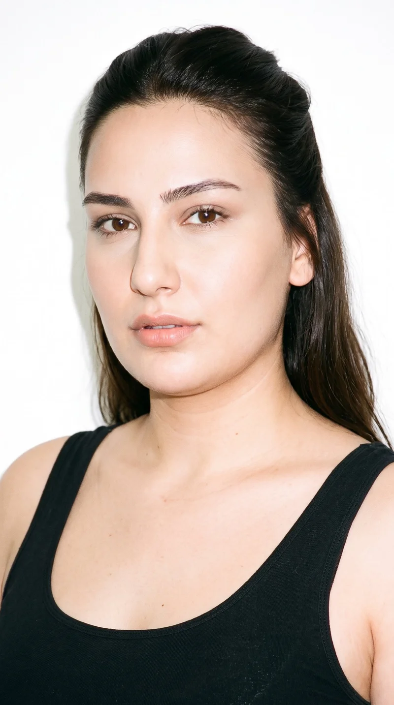 A 24-year-old Turkish woman with a medium olive skin tone and greenish-golden undertone has a long face with a tall forehead and elongated cheeks. Her eyebrows are medium in thickness, and her eyes are brown. She has medium lips and dark-brown hair styled with the upper section pulled back while the lower lengths hang loose.