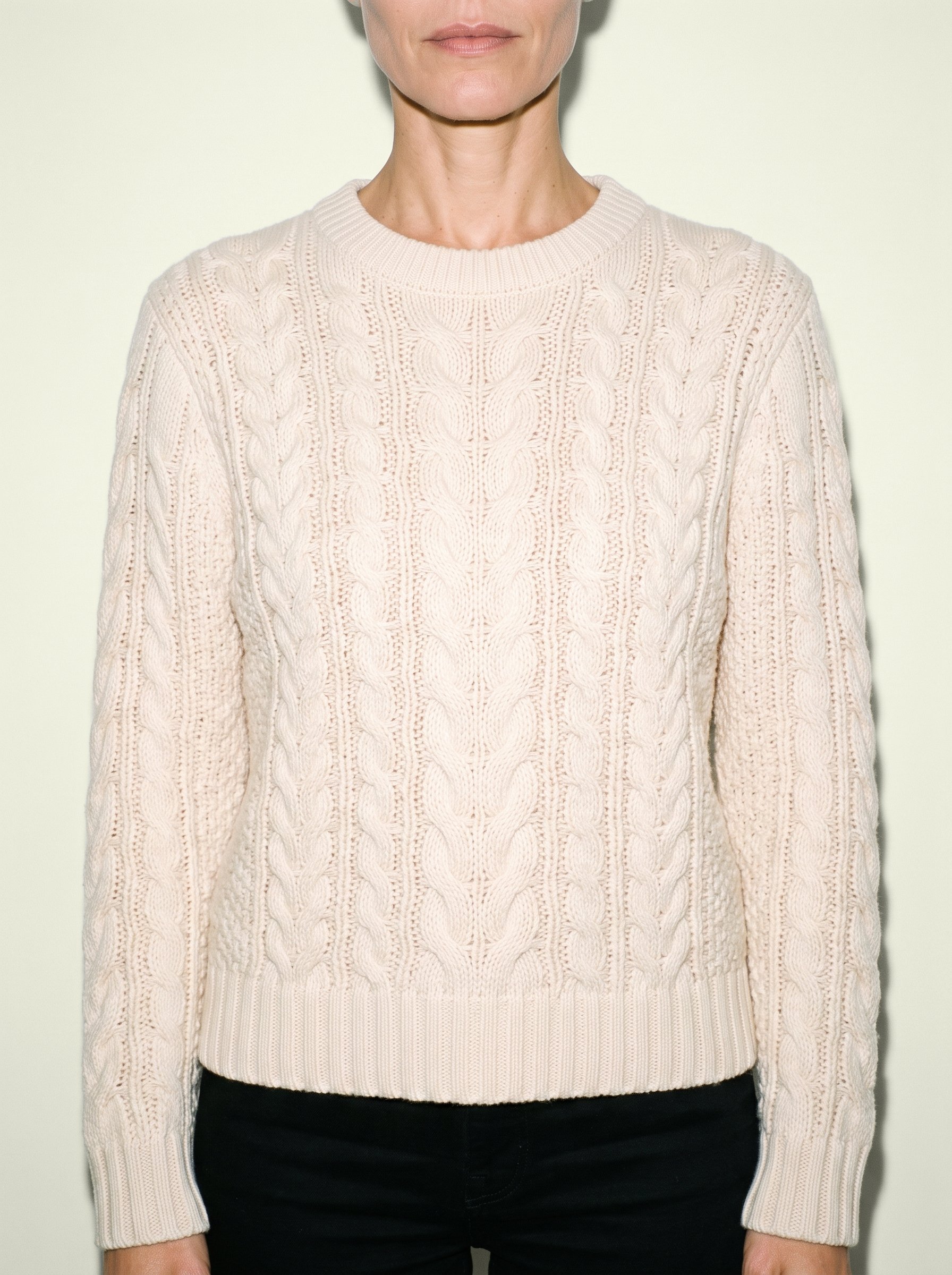 A woman's cream cable-knit pullover jumper shown from the front in a torso-framed view, with her arms relaxed. An ivory lace studio-wall backdrop is visible behind the jumper.