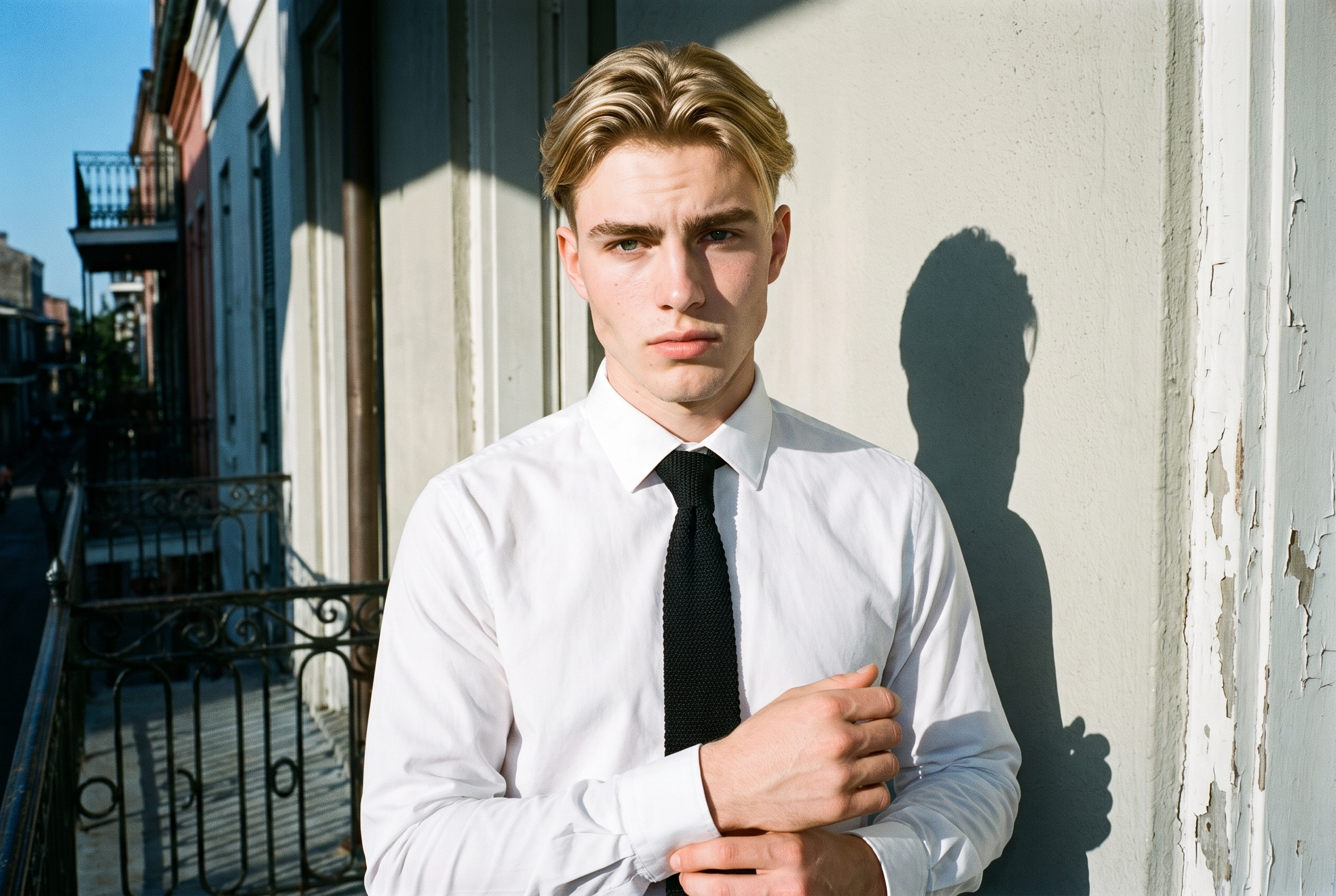 A 19-year-old Caucasian man is shown from the front in an upper-body view wearing a black knitted tie. He is touching his wrist with a curious expression, with a New Orleans balcony as the backdrop.