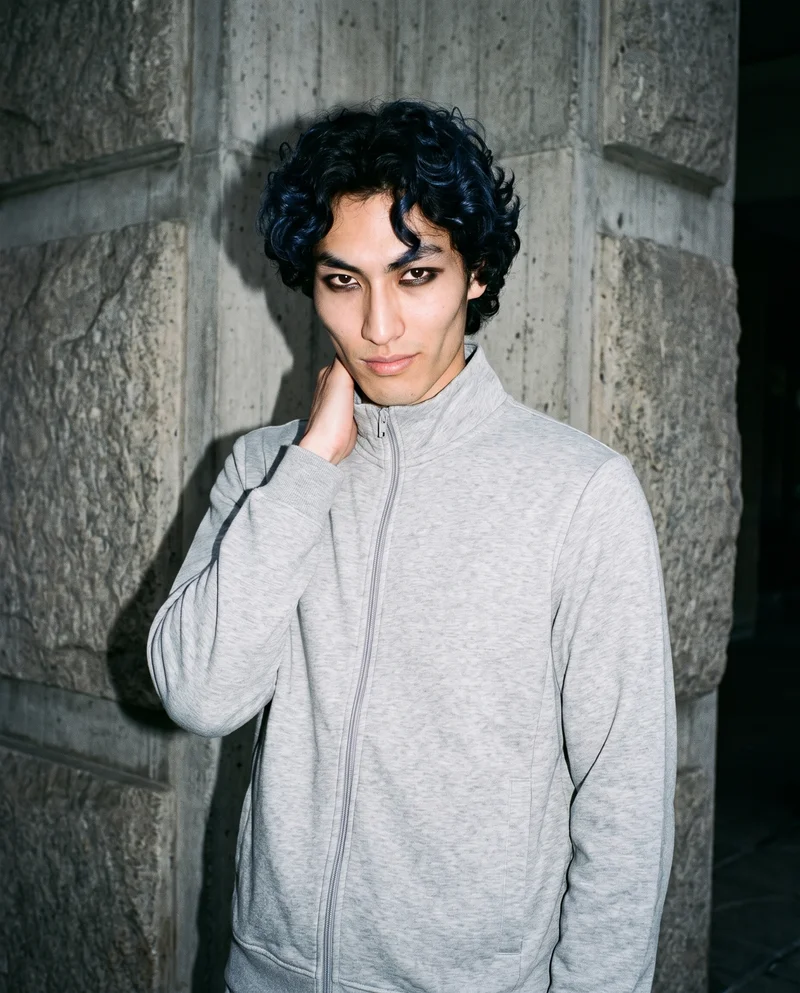 A 22-year-old Chinese man wears a grey marl tracksuit in a bust-framed front three-quarter view, with one hand resting on his neck and a natural facial expression. A Brutalist Monument Plaza forms the backdrop.