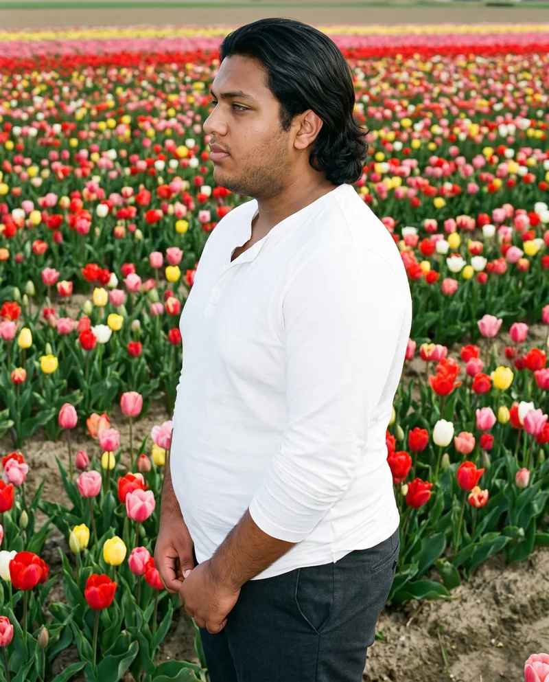 A 22-year-old Arab man wears a white Henley top in an upper-body side-view composition, with his hands held in front and a natural facial expression. A tulip field forms the backdrop.