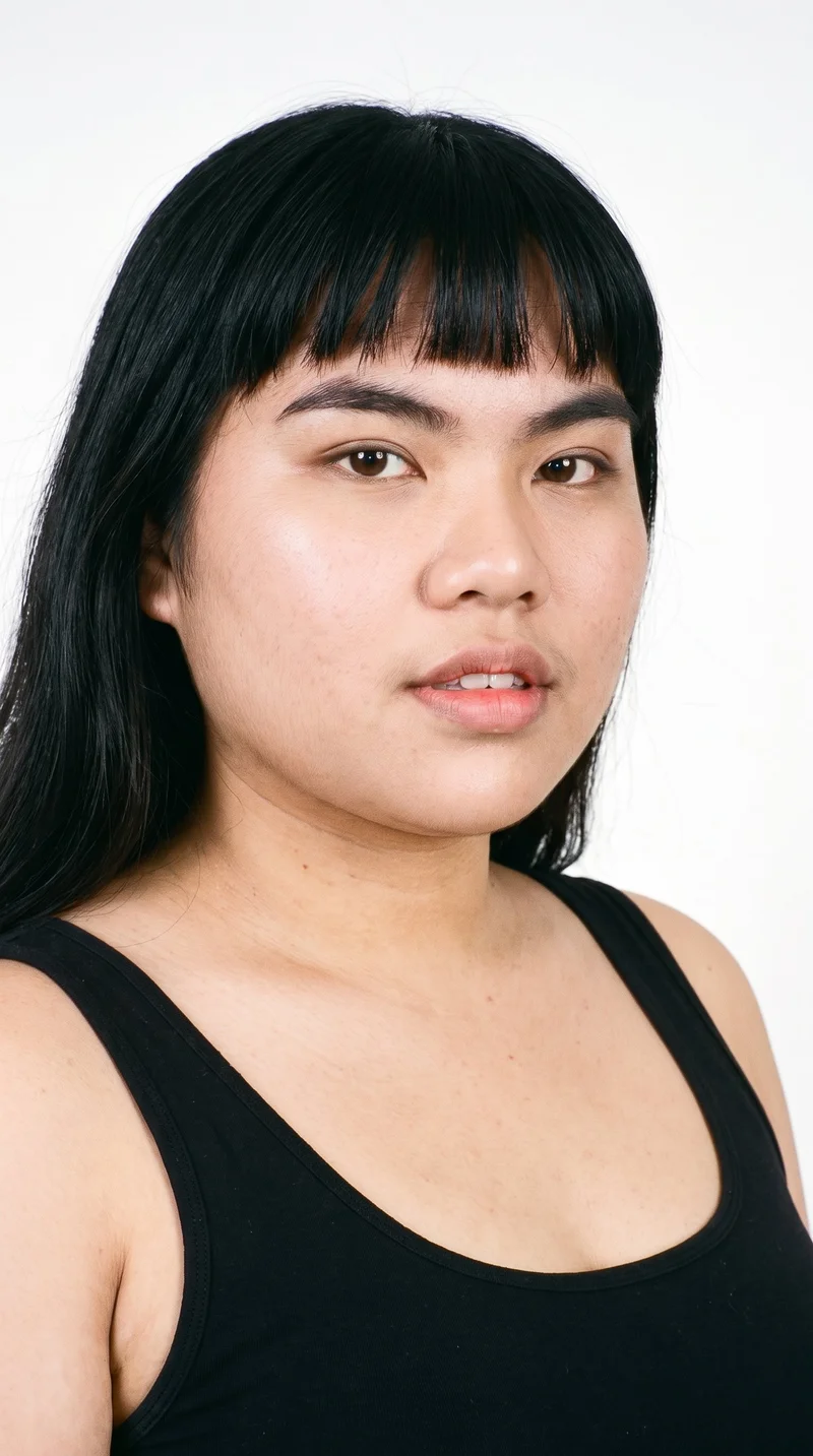 This 21-year-old Southeast Asian woman has lightly tanned fair skin with a neutral undertone. Her round face has soft, full contours with gentle curves and few angles. She has thick eyebrows, brown eyes, and very full lips. Black hair falls across her forehead in bangs, framing the upper face.
