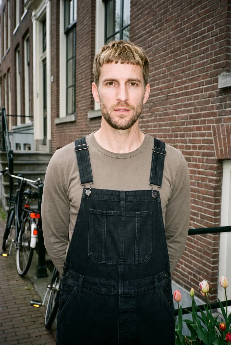 A 43-year-old Caucasian male model wears black denim dungarees in an upper-body view from a front three-quarter angle, with his hands behind his back and a natural facial expression. An Amsterdam canal house serves as the backdrop.