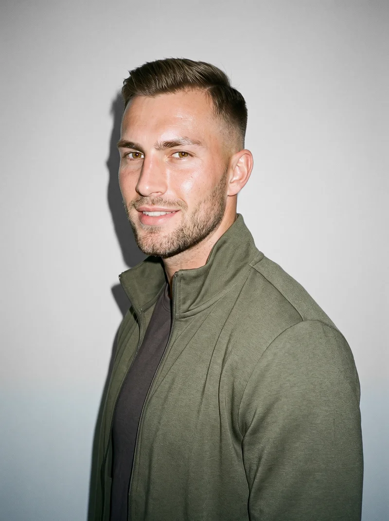 A 22-year-old Caucasian man wears a men's olive tracksuit in a bust-length view from a front three-quarter angle. He is smiling with his chin lowered, against a Seasalt studio-wall backdrop.