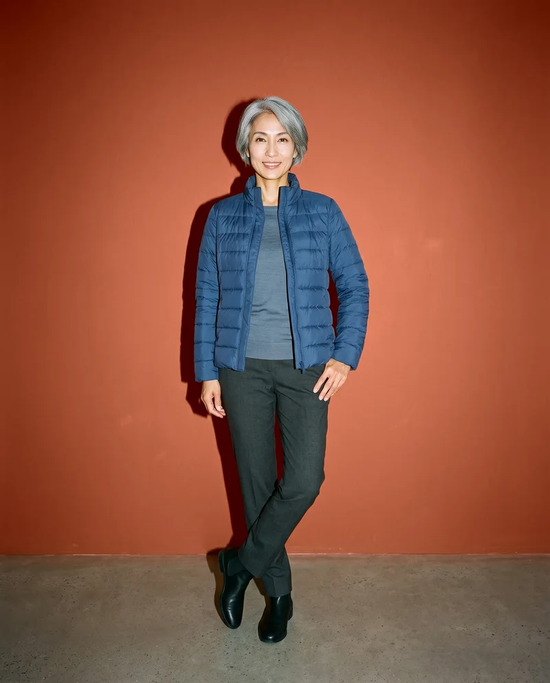 A 37-year-old Japanese woman wears a navy padded jacket in a front-facing environmental view, standing with one hip angled outward and smiling. A terracotta clay studio-wall backdrop surrounds her.
