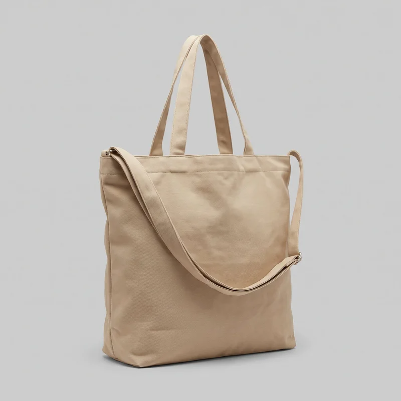 A beige rectangular canvas tote bag is shown in a standalone three-quarter view, with its flat front panel, open top, and structured boxy base visible. Twin upright carry handles rise from the top, while a detachable or adjustable shoulder strap connects at the sides with metal ring hardware. The woven canvas exterior has bound edges, a horizontal seam band below the opening, vertical side seams, and squared bottom corners.