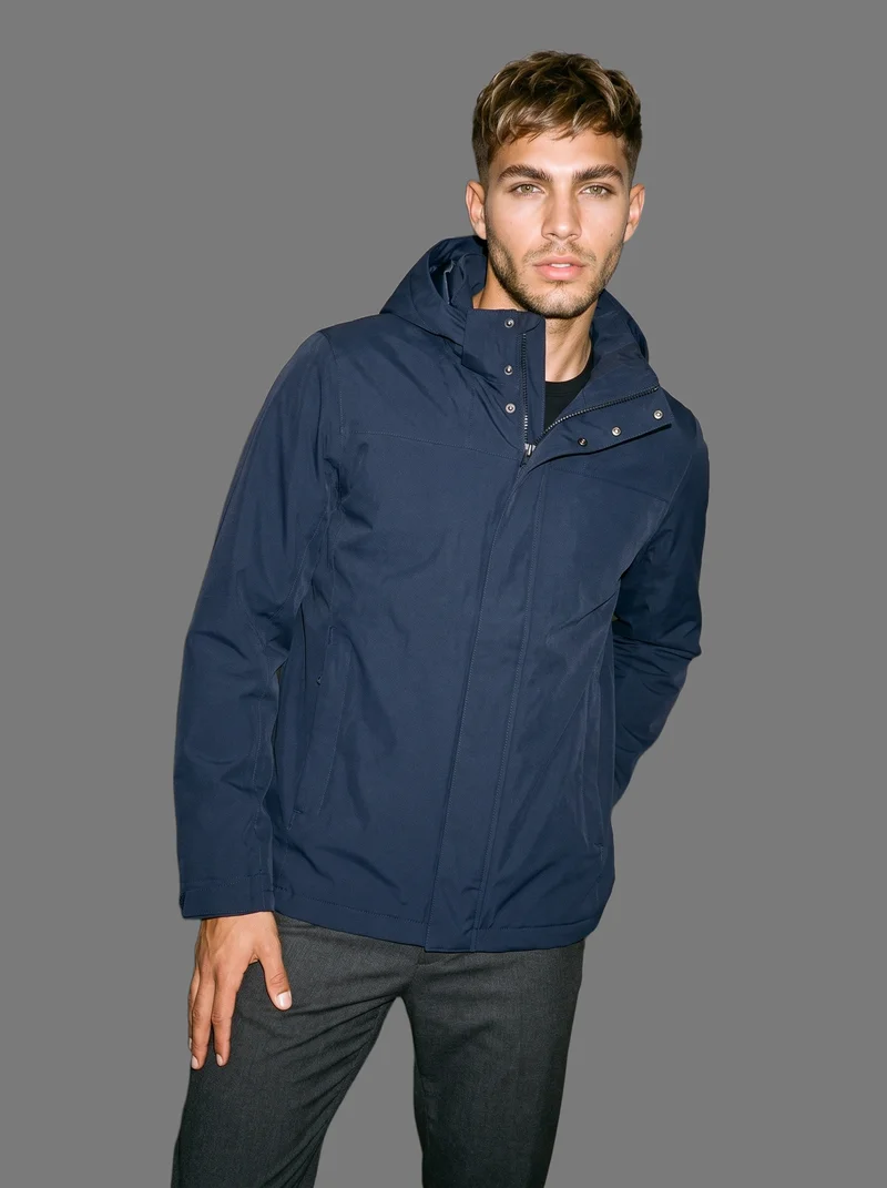 A 23-year-old Hispanic man wears a men's navy waterproof parka in a three-quarter-length front view, shown with a waist twist and a neutral expression. The image uses a flat-color Boulder backdrop.