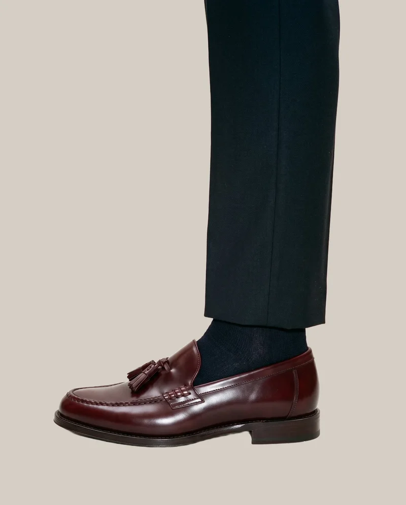 A men's burgundy tassel loafer shown from the side with the foot flat, cropped at the ankle against a London Fog flat-color backdrop.
