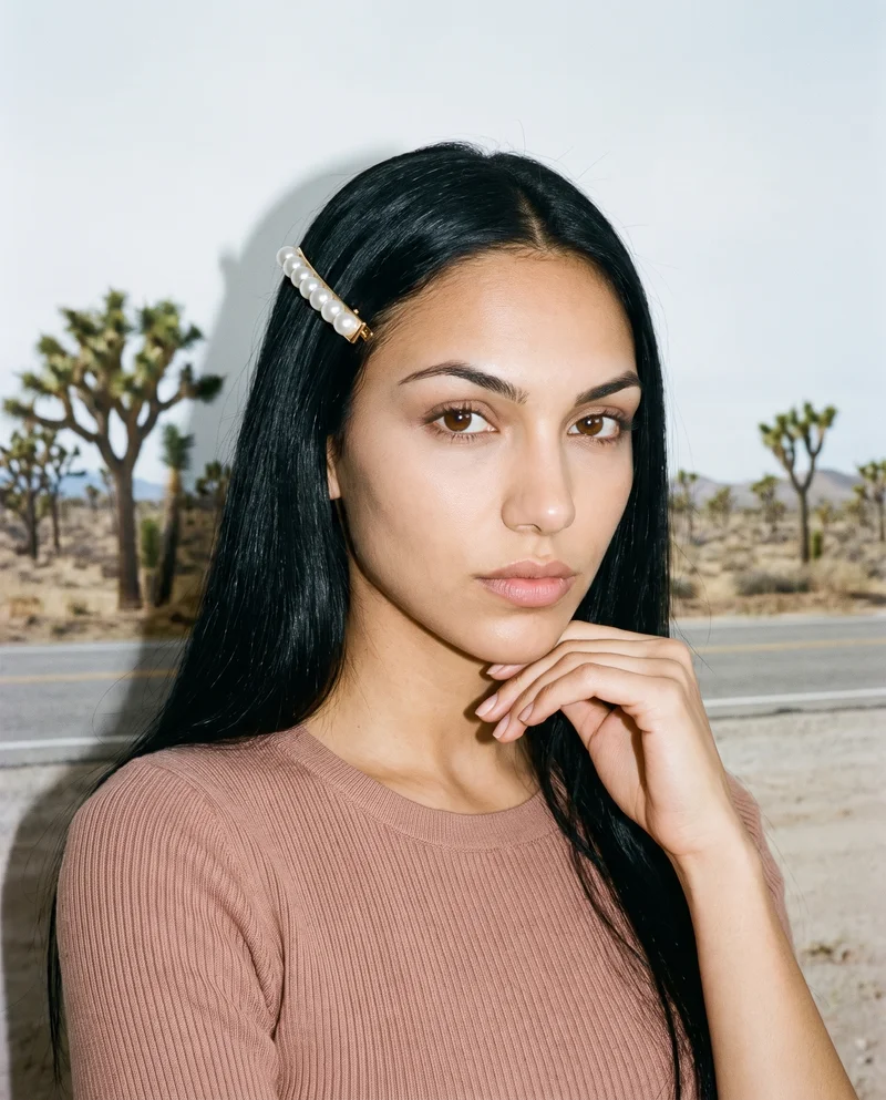 A 21-year-old Caucasian woman is shown from the bust up in a front view, wearing a pearl hair clip. Her hand rests at her jaw, and she has a confident expression. A Joshua Tree desert highway forms the backdrop.