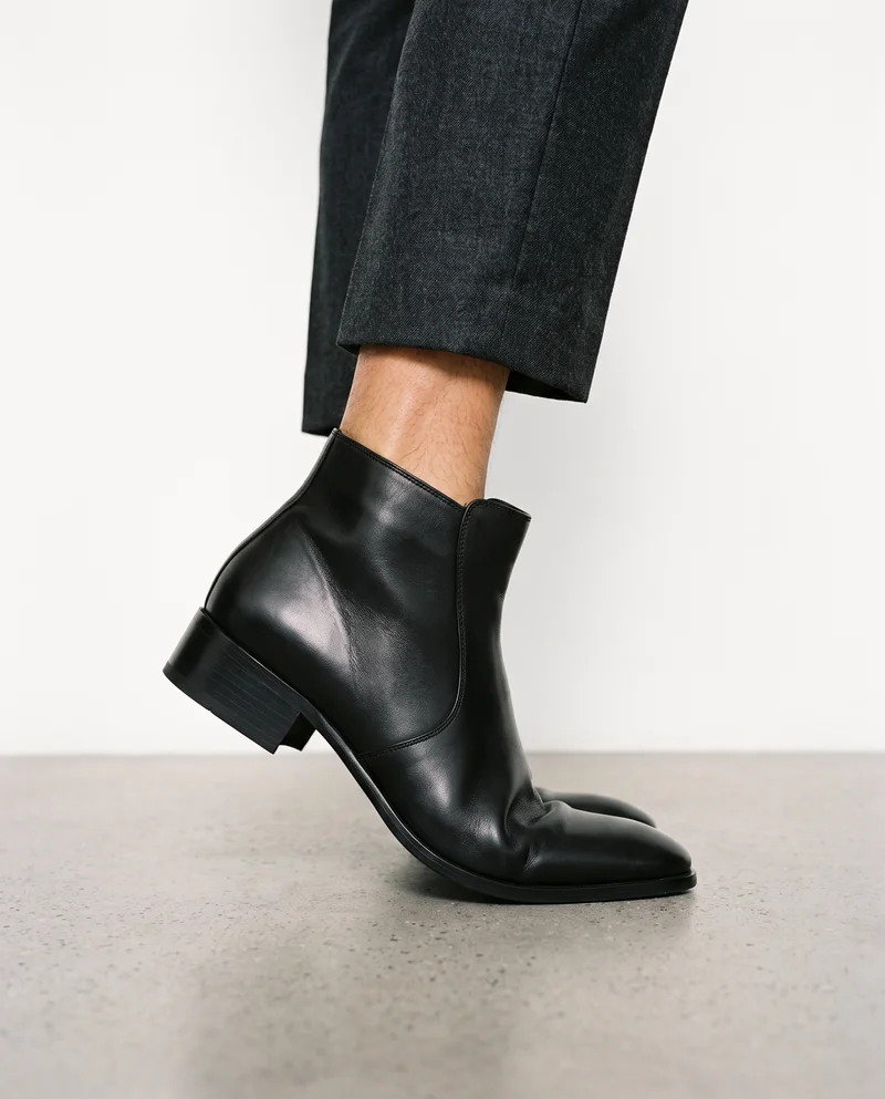 Men's heeled ankle boots shown in an ankle-framed side view, with the foot positioned on tiptoe against a Chantilly Lace studio-wall backdrop.