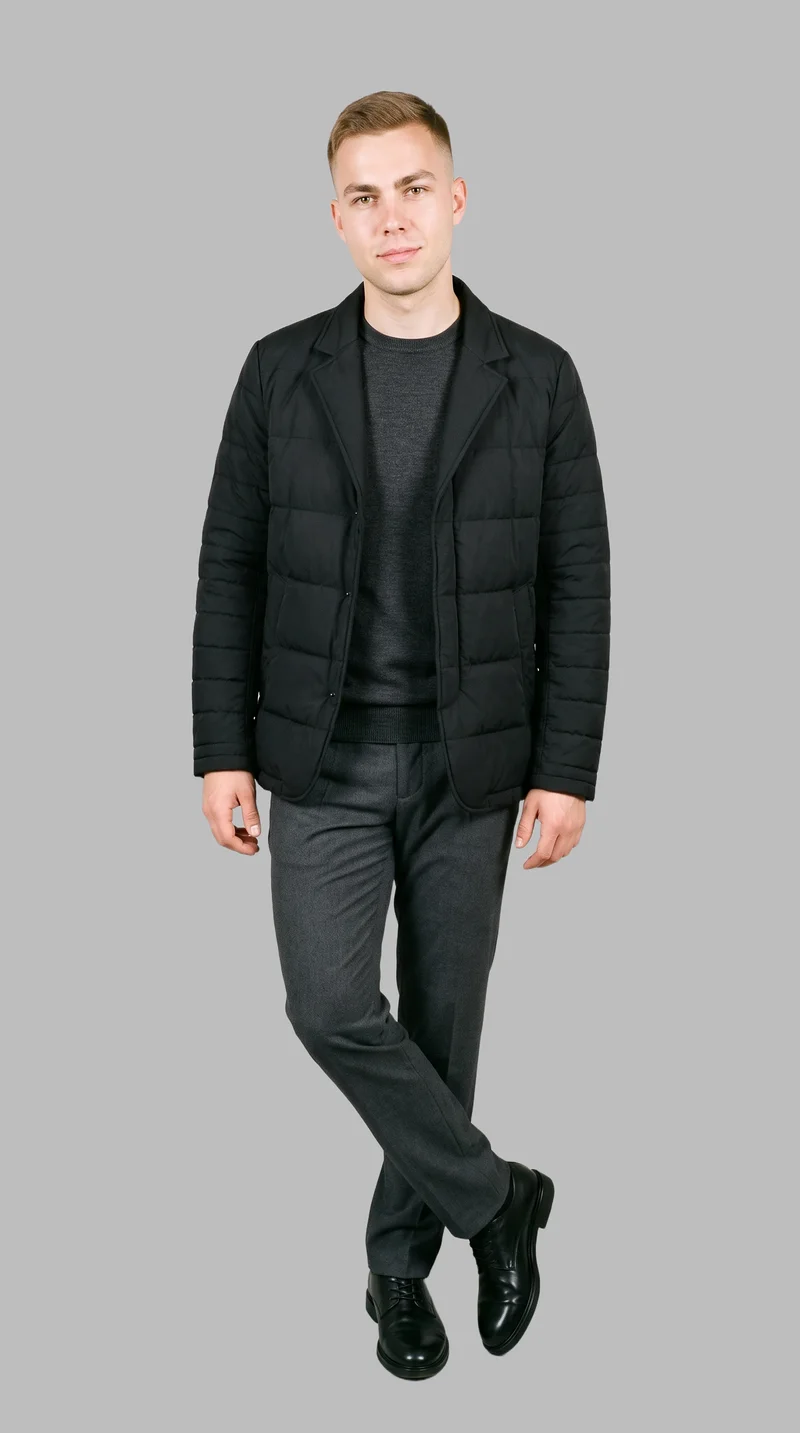 A 25-year-old Caucasian man wears a black puffer jacket in a full-body front three-quarter view. He stands with his ankles crossed against a dove grey backdrop, with a natural facial expression.