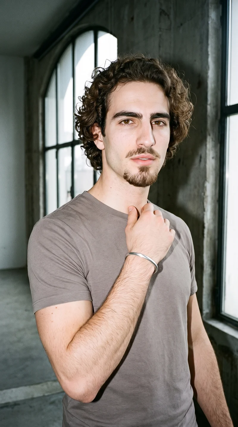 A 26-year-old Caucasian man is shown in an upper-body front three-quarter view with a soft expression and one hand at his collarbone, displaying a men's silver cuff bracelet. Massive loft windows with a golden tone form the backdrop.