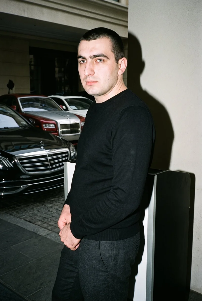 A 29-year-old Slavic male model wears a black pullover jumper in a three-quarter-length, angled-front view with his hands held in front. The image uses a valet stand backdrop.