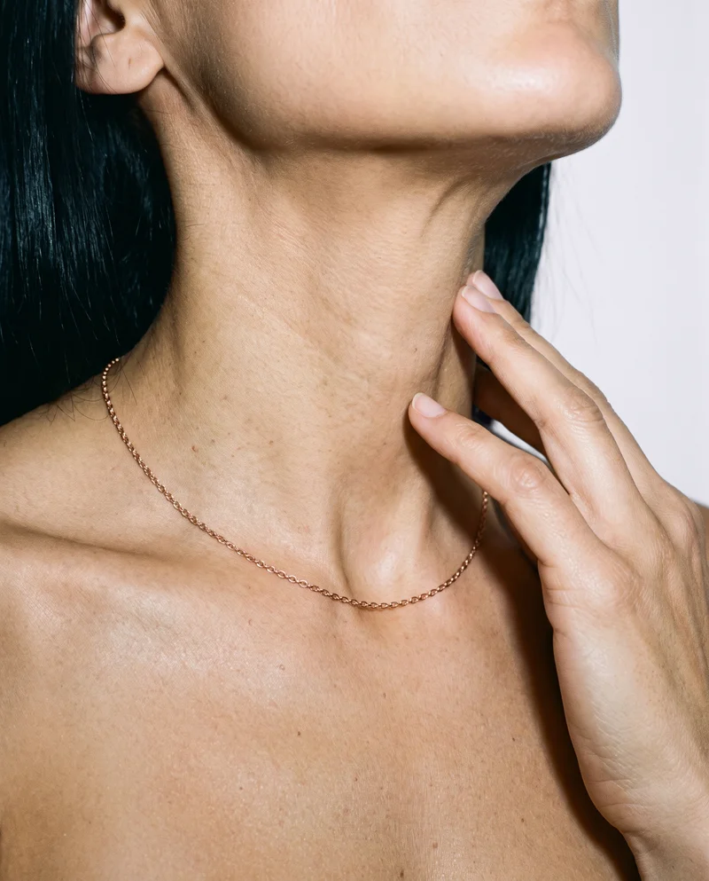 A woman's rose gold chain is shown around the neck in a front view, with fingers resting down the throat. The porcelain studio-wall backdrop frames the jewelry.