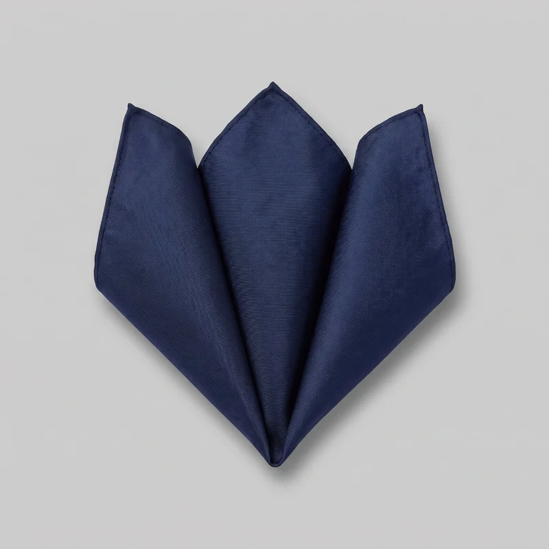 A navy pocket square is shown as a standalone accessory, pre-folded into a fan-like arrangement with three upright triangular points and a single downward V-shaped point. Overlapping diagonal folds create crisp panels, visible side seams, and pointed corners across the fine plain woven surface. The full folded silhouette is visible against a simple backdrop, with no logos, prints, patches, embroidery, or embellishments visible.