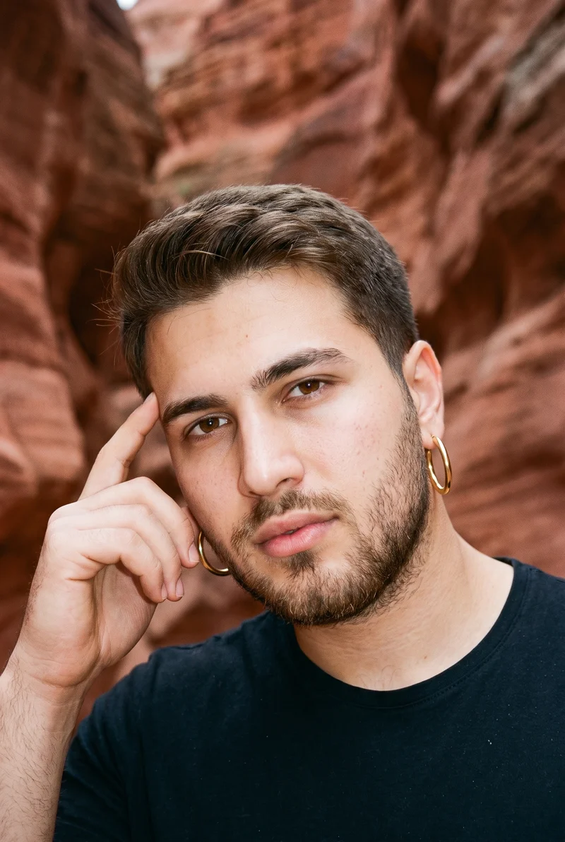A 20-year-old Turkish man is shown from the head in a front three-quarter view, wearing large gold hoop earrings. One finger rests at his temple, and he has a confident expression. Red rock formations appear as the backdrop.
