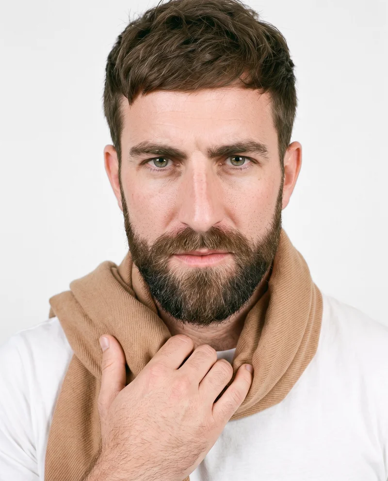 A 36-year-old Caucasian man is shown in a front-facing bust view wearing a camel cashmere scarf, with one hand at his collarbone and an intense expression. A cloud-white studio wall forms the backdrop.